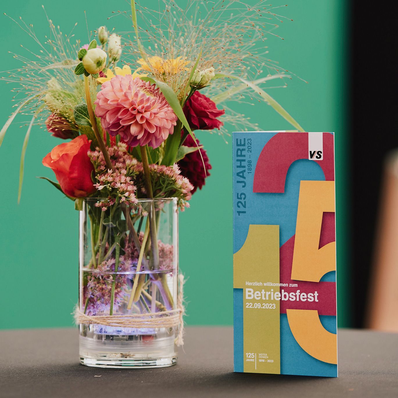The colourful invitation card for the VS 125th anniversary celebration stands next to a bouquet of flowers.