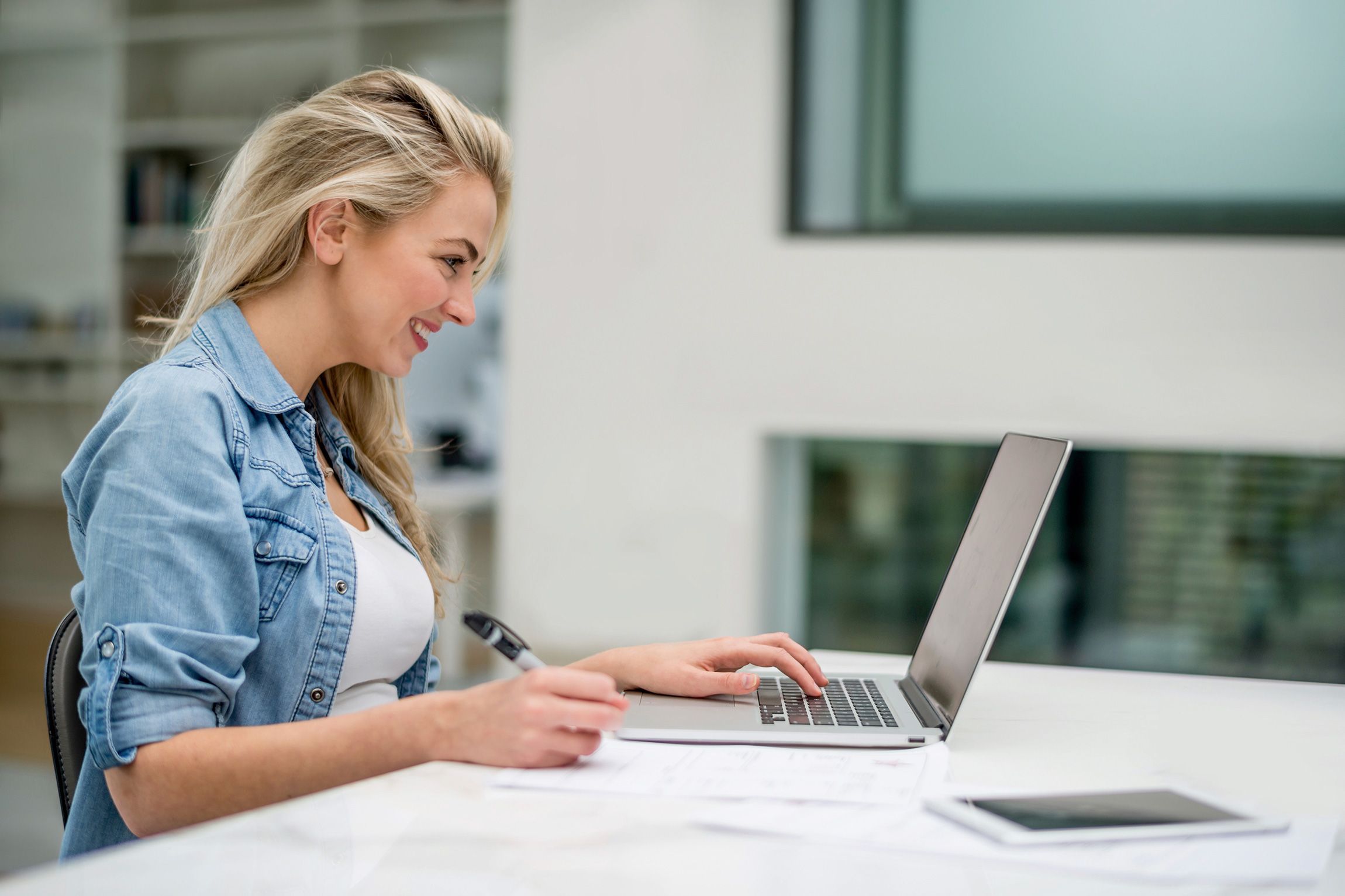 A woman sits at a laptop and writes something down on a piece of paper