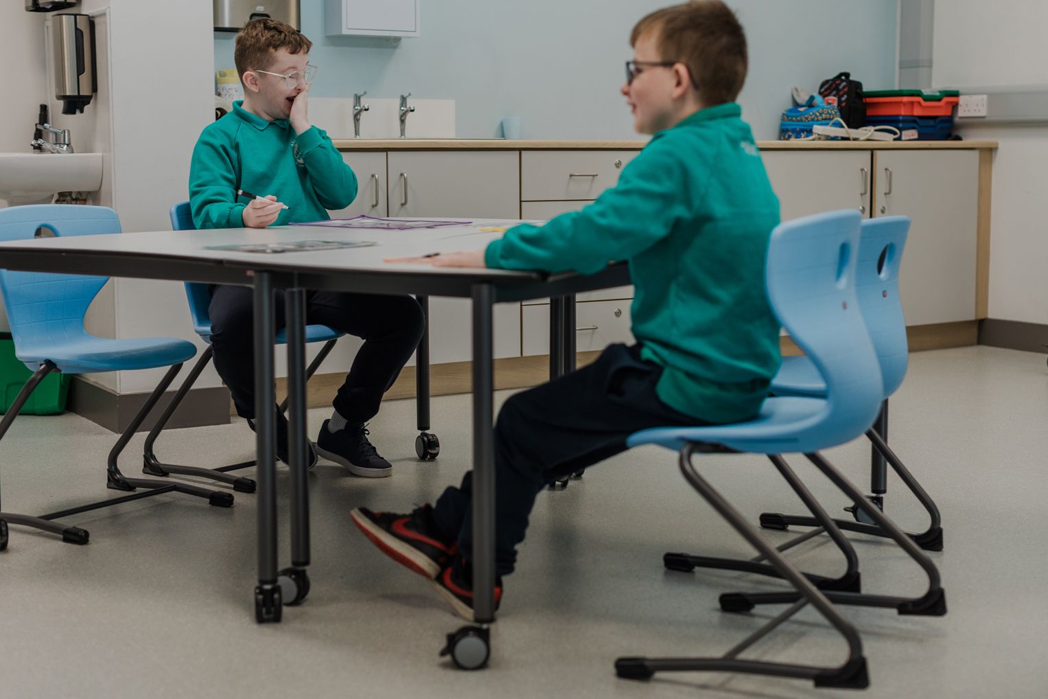 Zwei Schüler der Ardnashee School arbeiten an einem fahrbaren Tisch auf Rollen und sitzen dabei auf hellblauen Schwingstühlen.